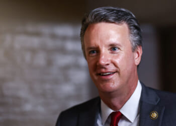 Smiling man in a suit and red tie stands indoors with a blurred, neutral background. He has short gray hair and wears a pin on his lapel.