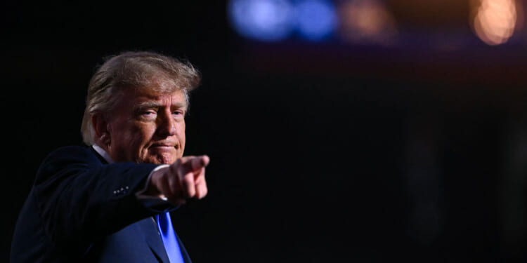 Republican President Donald Trump points forward with a serious expression, standing against a dark background with blurred lights in the upper right corner.