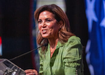 A woman in a green blouse speaks at a podium with a microphone. The background features a large, partially visible American flag with blue and white stars.