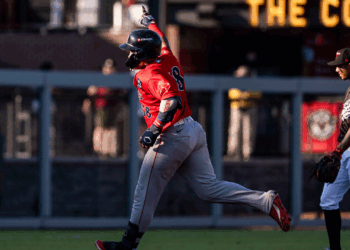 A baseball player in red celebrates with his arm raised while running, as another player in black and white stands nearby watching on the baseball field.