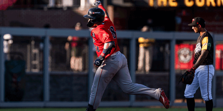 A baseball player in red celebrates with his arm raised while running, as another player in black and white stands nearby watching on the baseball field.