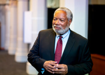 An older man with gray hair and a beard, wearing a dark suit, light blue shirt, and red polka dot tie, stands indoors, smiling with his hands clasped.