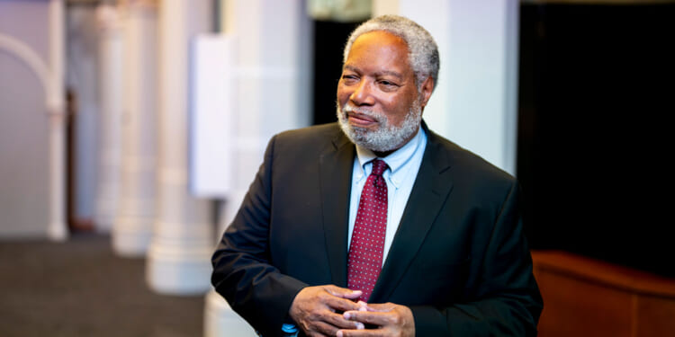 An older man with gray hair and a beard, wearing a dark suit, light blue shirt, and red polka dot tie, stands indoors, smiling with his hands clasped.