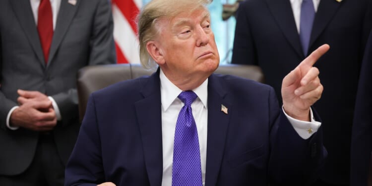 A man in a navy suit and purple tie sits at a desk, pointing to his right while others stand behind him. An American flag is visible in the background.