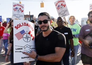 A group of people march holding Solidarity signs showing U.S. and Canadian flags, a compass, and shaking hands. The crowd appears diverse and determined, participating in a labor support event on a city street.