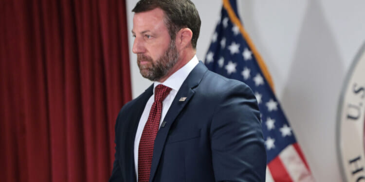 A man in a navy suit and red tie stands in front of an American flag and a red curtain, appearing serious and focused.
