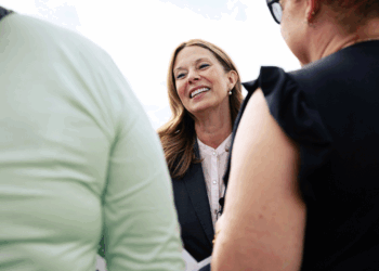 Amy Actonin a blazer smiles while talking to two people outdoors. The image is bright, and the mood appears friendly and engaging.