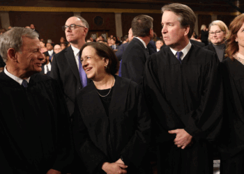 Five judges in black robes stand together in a formal setting, engaged in conversation. Several other people are visible in the background, most seated and dressed formally.