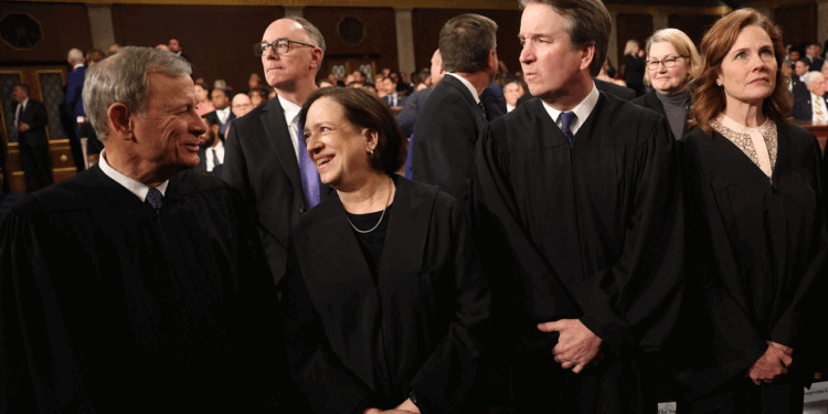 Five judges in black robes stand together in a formal setting, engaged in conversation. Several other people are visible in the background, most seated and dressed formally.