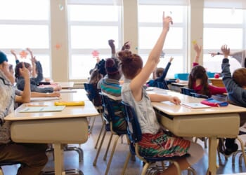 Kids in a classroom seated at their desks are raising their hands.