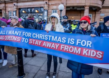 A group of people stand outside holding a large banner reading, All I want for Christmas is for drag queens to stay away from kids. Police and bystanders are visible in the background.