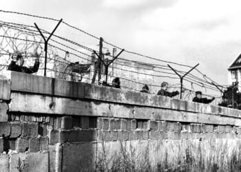 Black-and-white photo of people peering over a concrete wall topped with barbed wire. A building is visible in the background, and grass grows along the base of the wall.