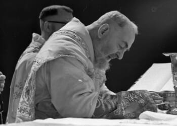 Padre Pio, a bearded priest in ornate vestments leans forward in prayer at an altar, with a chalice and open book in front of him, another clergy member stands nearby.