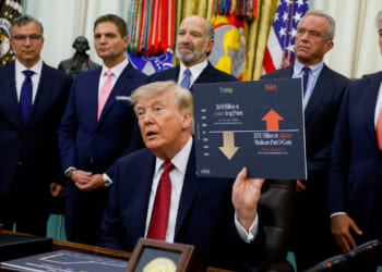 Donald Trump sits at a desk holding a chart comparing drug prices, surrounded by six men in suits, with U.S. flags and military emblems in the background, highlighting his focus on the economy.