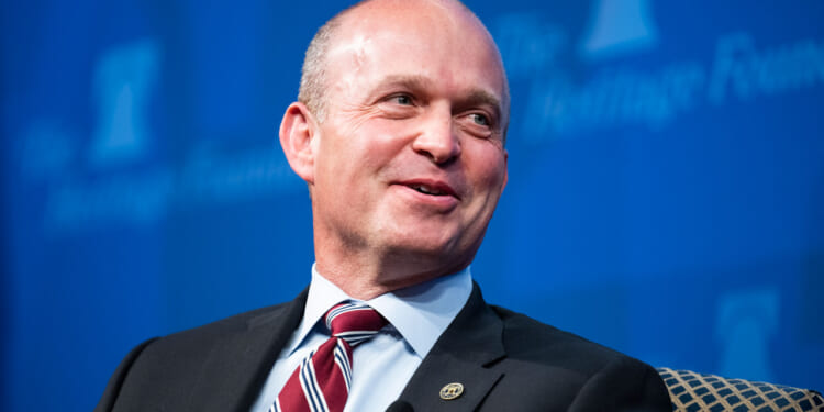 Kevin Roberts in a suit and striped tie smiles while seated against a blue backdrop with partially visible text.