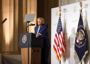 A man in a suit stands at a podium with the presidential seal, flanked by two US flags and banners, delivering a speech in a formal setting.