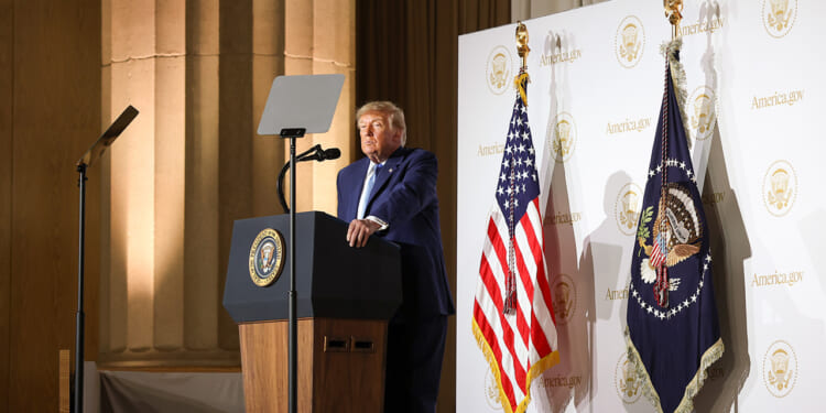 A man in a suit stands at a podium with the presidential seal, flanked by two US flags and banners, delivering a speech in a formal setting.