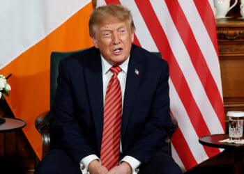 A man in a navy suit with a red striped tie sits between Irish and American flags, with flowers, a glass of water, and teapots on a wooden table beside him.