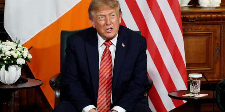 A man in a navy suit with a red striped tie sits between Irish and American flags, with flowers, a glass of water, and teapots on a wooden table beside him.