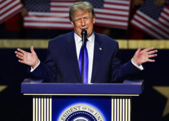 Donald Trump in a blue suit speaks at a podium with the presidential seal, gesturing with both hands, while American flags are held in the background.