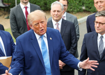A man in a blue suit speaks animatedly to a group of men in suits, who stand attentively behind him outdoors.