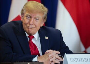 A man in a navy suit and red tie sits at a table with a sign labeled United States, in front of international flags, smiling slightly and looking forward.