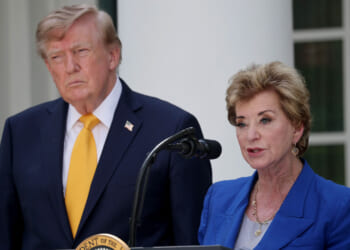 A man in a suit and yellow tie stands beside a woman speaking at a podium outdoors. The woman wears a blue jacket and stands in front of a microphone. Both appear serious.