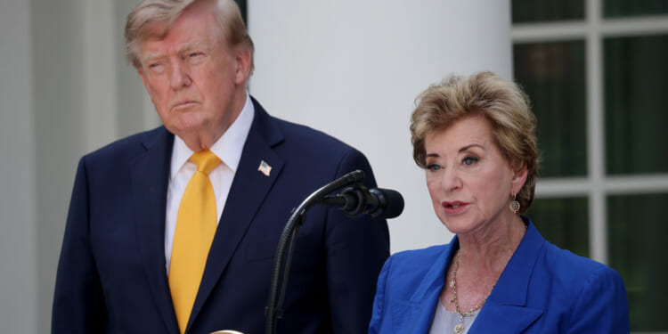 A man in a suit and yellow tie stands beside a woman speaking at a podium outdoors. The woman wears a blue jacket and stands in front of a microphone. Both appear serious.