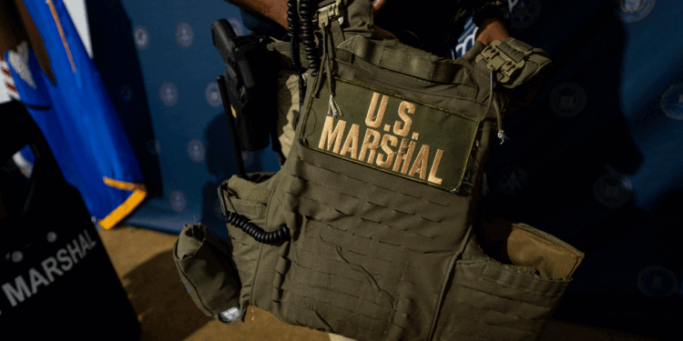 A person holds a tactical vest labeled U.S. Marshal in large yellow letters, with a radio attached. A U.S. Marshals shield and blue flag are visible in the background.