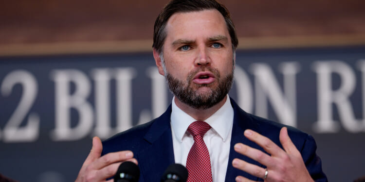 A man in a suit and red tie speaks passionately at a podium with microphones, gesturing with his hands. The blurred background shows partial text on a sign.
