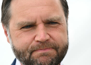 A close-up of JD Vance with short brown hair, a full beard, and a serious expression, with a blurred light background.