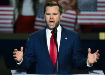 A man in a blue suit and red tie speaks passionately at a podium with a microphone; blurred people behind him hold American flags.