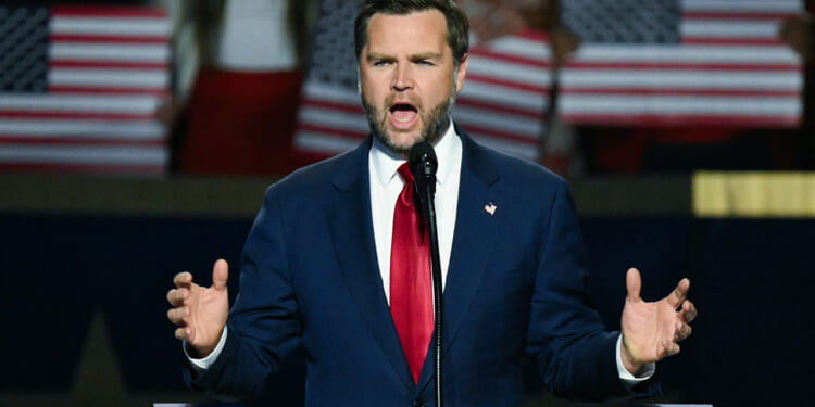 A man in a blue suit and red tie speaks passionately at a podium with a microphone; blurred people behind him hold American flags.