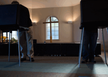 Two people stand at separate voting booths in a room with carpeted floors, sunlight streaming in, and a cross and windows visible in the background.