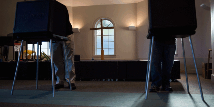 Two people stand at separate voting booths in a room with carpeted floors, sunlight streaming in, and a cross and windows visible in the background.