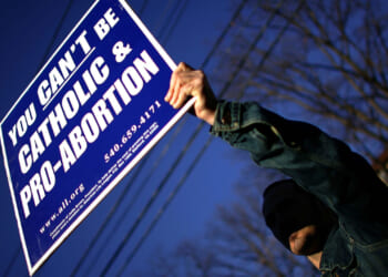 A person holds a large sign reading, You cant be Catholic & pro-abortion, with contact information and a website, against a blue sky and some tree branches.