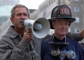 A man in a jacket speaks through a megaphone while standing with his arm around a firefighter wearing a helmet marked 164 and a respirator around his neck. Buildings are visible in the background.