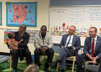 Four HHS officials sit in a classroom, one holding a dog. Behind them are colorful bulletin boards, childrens art, and classroom instructions displayed on the wall.