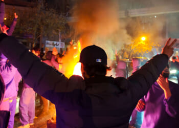 A person with arms raised faces a crowd of Iranian people gathered around a fire at night, with smoke and orange-yellow flames lighting the scene. Other people are visible, some with hands raised, in a city street environment.