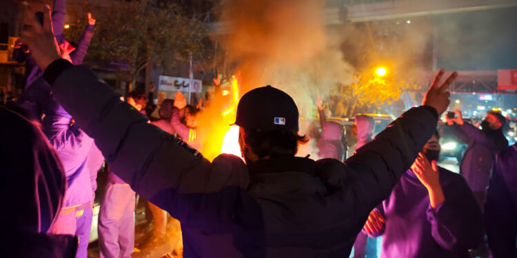 A person with arms raised faces a crowd of Iranian people gathered around a fire at night, with smoke and orange-yellow flames lighting the scene. Other people are visible, some with hands raised, in a city street environment.