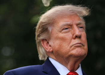 President Donald Trump, a man with light skin and blond hair wearing a navy suit, white shirt, and red tie looks upwards with a serious expression. The background is blurred greenery.