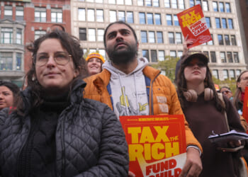 A diverse group of people at a rally hold signs reading TAX THE RICH, FUND CHILD CARE with tall city buildings in the background. The crowd appears attentive and focused on a speaker or event ahead.