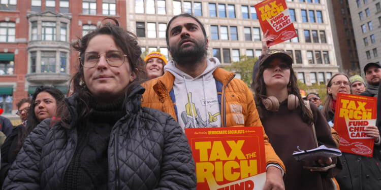 A diverse group of people at a rally hold signs reading TAX THE RICH, FUND CHILD CARE with tall city buildings in the background. The crowd appears attentive and focused on a speaker or event ahead.