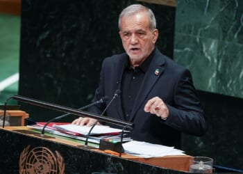 A man in a dark suit speaks at a podium with the United Nations emblem, addressing an audience. Papers and a microphone are on the podium, and a marble wall is in the background.