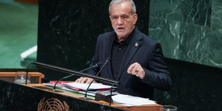 A man in a dark suit speaks at a podium with the United Nations emblem, addressing an audience. Papers and a microphone are on the podium, and a marble wall is in the background.