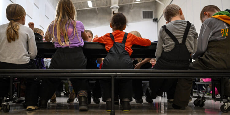 A group of children wearing winter clothes sit closely together on benches at a table in a brightly lit room, seen from behind.