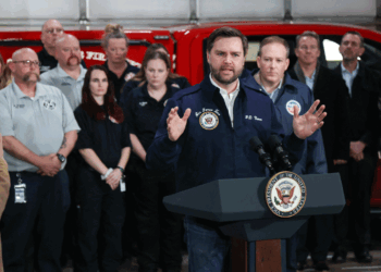 JD Vance speaks at a podium labeled Vice President of the United States while a group of people, some in uniforms, stand behind him in front of a red emergency vehicle.
