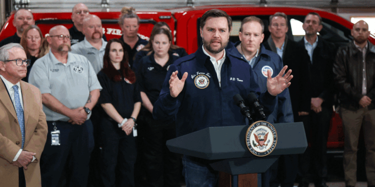 JD Vance speaks at a podium labeled Vice President of the United States while a group of people, some in uniforms, stand behind him in front of a red emergency vehicle.