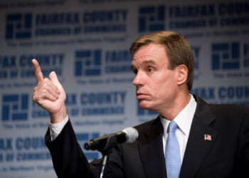 Mark Warner in a suit gestures with his right hand while speaking at a podium. The background displays the Fairfax County Chamber of Commerce logo and text repeatedly.