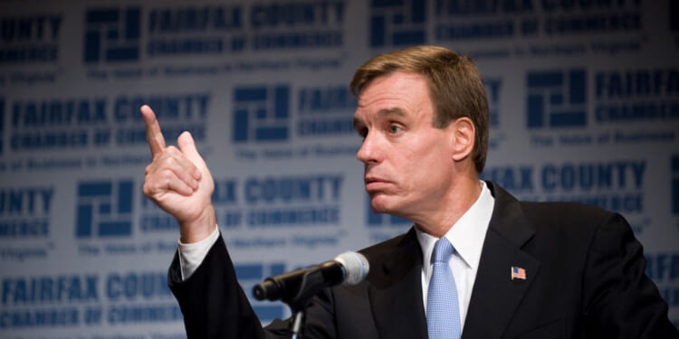 Mark Warner in a suit gestures with his right hand while speaking at a podium. The background displays the Fairfax County Chamber of Commerce logo and text repeatedly.